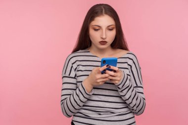Cep telefonunda yazan, blog okuyan, sosyal ağda sohbet eden, çevrimiçi iletişim için mobil uygulama kullanan ciddi özenli genç kadının portresi. Pembe arka planda kapalı stüdyo çekimleri