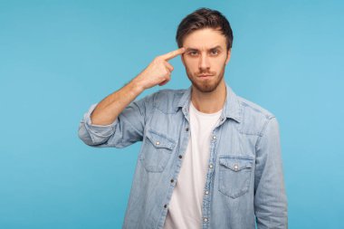 Crazy idea! Portrait of man in denim shirt showing stupid gesture, looking at camera with condemnation and blaming for insane plan, dumb suggestion. indoor studio shot isolated on blue background
