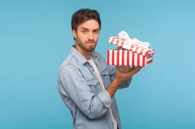 Portrait of frustrated man in denim shirt holding opened gift box and looking at camera with disappointed expression, unpacking present, dissatisfied with awful birthday surprise. indoor studio shot clipart