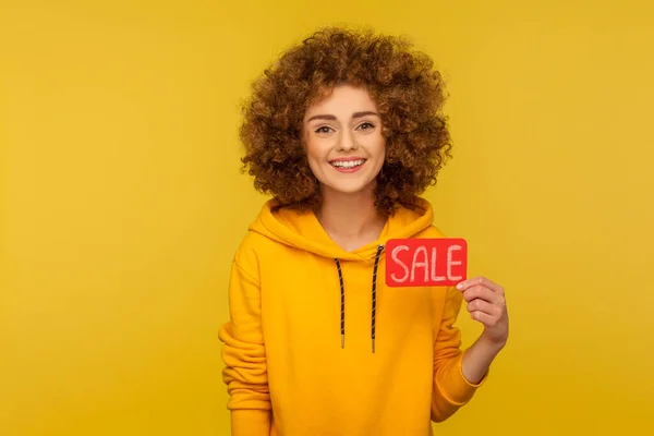 Shopping discount, low prices. Portrait of happy curly-haired woman in urban style hoodie showing Sale inscription and looking with toothy smile. indoor studio shot isolated on yellow background
