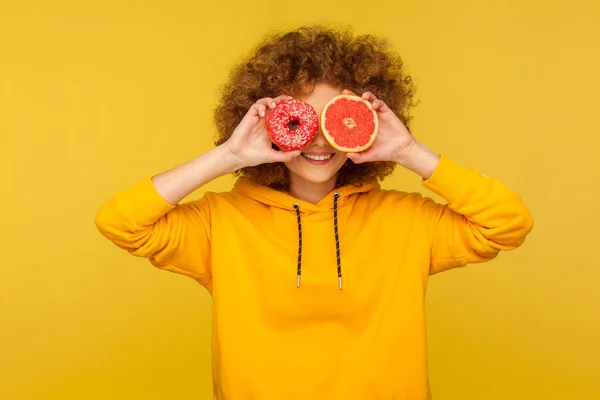 建议的甜点对水果 一个快乐的卷发女人的画像 她穿着都市风格的帽衫 用新鲜的柚子和甜甜的甜甜圈 垃圾食品蒙住眼睛 在黄色背景下隔离的室内拍摄 — 图库照片