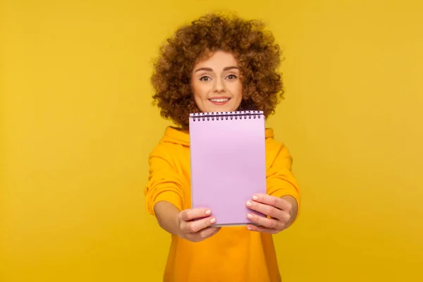 快乐快乐的学生的肖像 卷发女人穿着都市风格的帽衫 展示空白笔记本 在纸上做广告的区域 复制待办事项的清单 在黄色背景下隔离的演播室拍摄 — 图库照片