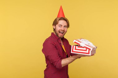 Kareli gömlekli ve parti konisi hediye kutusundaki heyecanlı adam şok olmuş bir ifadeyle kameraya bakıyor, doğum günü hediyesi paketini açıyor. Sarı arka planda izole edilmiş kapalı stüdyo çekimi