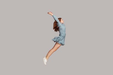 Side view girl in dress levitating hovering in mid-air with raised hands, model looking away concentrated focused, jumping trampoline gaining speed. indoor studio shot isolated on gray background clipart