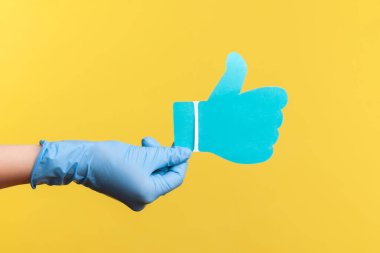 Profil yan görüntüsü, insan elinin mavi cerrahi eldivenlerle çekilmiş ve kağıt şeklindeki işaretleri gösteriyor. İçerde, stüdyoda, sarı arka planda izole edilmiş..