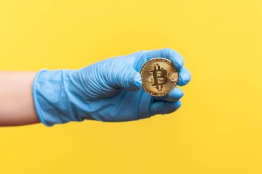 Profil yan görüntüsü, elinde bitcoin sembolü olan mavi eldivenli insan eli. İçerde, stüdyoda, sarı arka planda izole edilmiş..