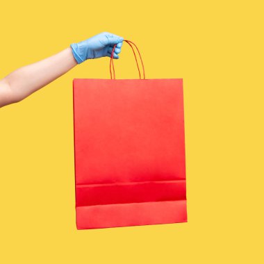 Kırmızı alışveriş torbası tutan mavi eldivenli insan elinin profil yan görüntüsü. İçerde, stüdyoda, sarı arka planda izole edilmiş..