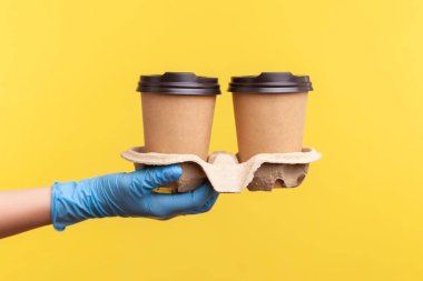 Profil yan görüntüsü. Mavi eldivenli insan eli. Elinde sıcak yemek kupaları var. İçerde, stüdyoda, sarı arka planda izole edilmiş..