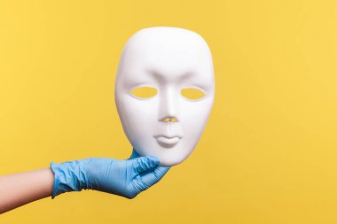 Profil yan görüntüsü: Mavi eldivenli insan eli beyaz maske modelini tutuyor. İçerde, stüdyoda, sarı arka planda izole edilmiş..
