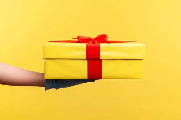 Stock image Profile side view closeup of human hand in blue surgical gloves holding yellow gift box. indoor, studio shot, isolated on yellow background.