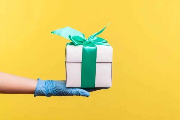 Profile side view closeup of human hand in blue surgical gloves holding white gift box. sharing, giving or delivery concept. indoor, studio shot, isolated on yellow background.