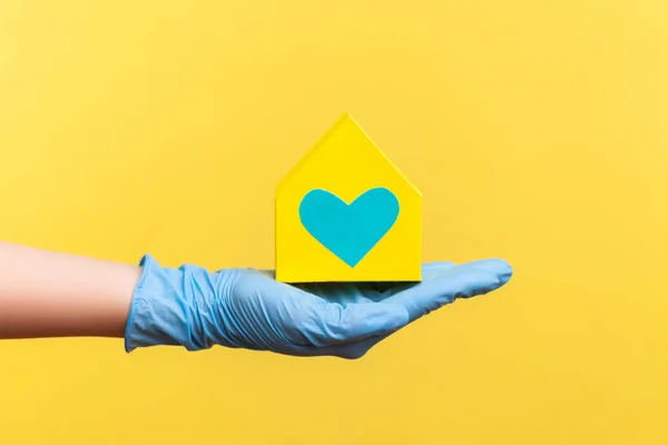 プロファイル側ビュー愛とケアと手に黄色の紙の家の外観を保持青い外科手袋で人間の手の閉鎖 黄色の背景に隔離された屋内スタジオでの撮影 — ストック写真