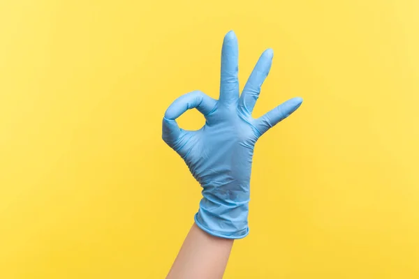 Profile side view closeup of human hand in blue surgical gloves showing Ok sign or number 3 with fingers. indoor, studio shot, isolated on yellow background.