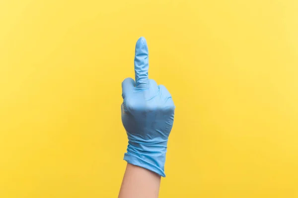 Profile side view closeup of human hand in blue surgical gloves showing middle finger. indoor, studio shot, isolated on yellow background.