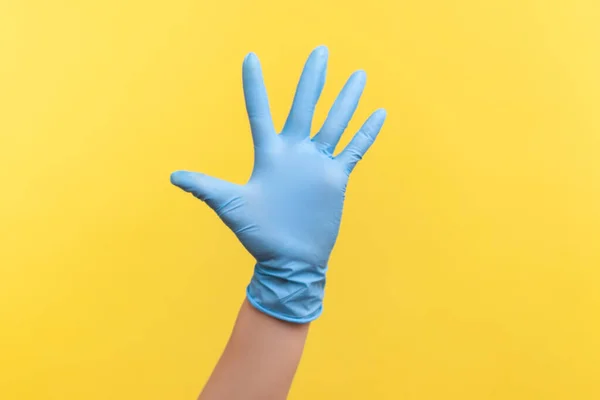 Profile side view closeup of human hand in blue surgical gloves showing number five with hand or waving hand to greeting. indoor, studio shot, isolated on yellow background.