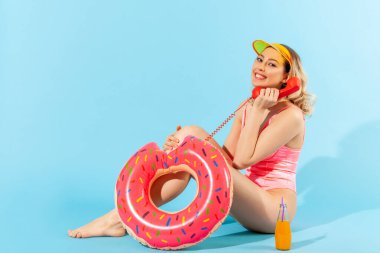 Uzun boylu, heyecanlı, mayolu bir kadın lastik yüzük, kokteyl ve telefon ahizesiyle oturuyor ve dişlek dolu bir gülümsemeyle kameraya bakıyor. Mavi arka planda kapalı stüdyo çekimleri