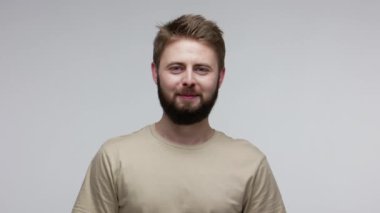 İyi huylu, genç ve sakallı bir adam kameraya bakıyor dişlek bir gülümsemeyle, hayatın tadını çıkarıyor, kaygısızca pozitif hissediyor ve gülüyor, iyimser bir bakış açısıyla. Gri arka planda kapalı stüdyo çekimi
