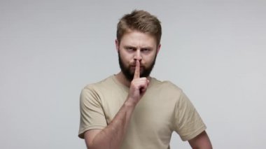 Shh, sessiz ol! Ciddi sakallı adam sessiz kalmak ya da gizlilik istiyor. Parmağını dudağına dayayıp jest yapıyor. Sakin ol ve gizli bilgileri söyleme. Gri arka planda kapalı stüdyo çekimi