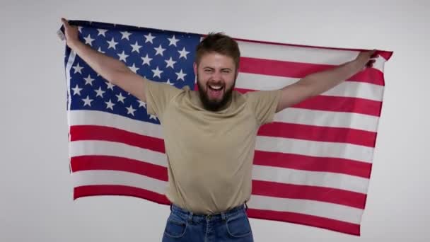 Patriotický radostný vousatý muž mávající americkou vlajkou, balící v USA prapor a křičící radostí, oslavující Den nezávislosti, svobodu lidských práv. vnitřní studio záběr izolované na šedém pozadí