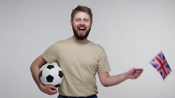 快乐的足球迷留着胡子 穿着T恤 拿着足球 挥动着英国国旗 为赢得进球而兴奋 为最喜欢的运动而欢呼 在灰色背景下隔离的室内拍摄 — 图库视频影像