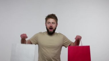Mağaza müşterisinin heyecanlı mutlu duyguları, tişörtlü, sakallı, alışveriş torbası kaldıran adam, satın alma, satış ve indirimden memnun görünüyor. Gri arka planda kapalı stüdyo çekimi