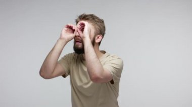 Meraklı, meraklı, sakallı bir adam uzak ufku gözetliyor dürbünle el hareketini izliyor ve kameraya bakarken şaşkınlığını dile getiriyor. Gri arka planda kapalı stüdyo çekimi