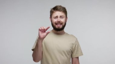 Genç sakallı adam hayal kırıklığına uğramış bir ifadeyle kameraya bakıyor ve parmaklarıyla biraz jest yapıyor, küçük boy, düşük ölçekli, daha fazlasına ihtiyacı var. Gri arka planda kapalı stüdyo çekimi
