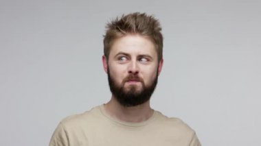 Optimistic funny bearded guy looking at camera cross eyed, showing tongue out, making stupid dumb face and aping, fooling around with brainless grimace. indoor studio shot isolated on gray background