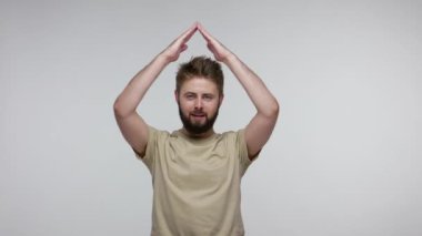 Sakallı hoş bir adam teras jestinin altında duruyor ve kameraya dişlek gülüşüyle bakıyor, kendi evini düşlüyor, hayatı ve güvenliği garanti altına alıyor, sigortası var. Gri arka planda kapalı stüdyo çekimi