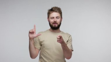 Pozitif sakallı adam L işareti ile duruyor, kaybeden hareketi gösteriyor ve kamerayı işaret ediyor, başarısızlıkla alay ediyor, şanssız bir gün, işten çıkarılma. Gri arka planda kapalı stüdyo çekimi