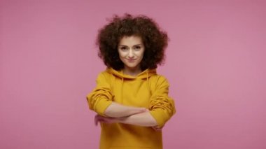 Kapüşonlu neşeli, mutlu afro saçlı kız ellerini çapraz yapıp kameraya dişlek bir gülümsemeyle bakıyor, iyimserliğini ve olumlu duygularını ifade ediyor. Stüdyo çekimi izole, pembe arkaplan