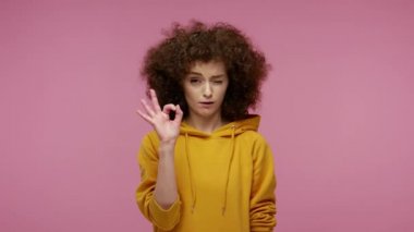 Ben iyiyim! Kapüşonlu afro saçlı oldukça olumlu bir kız el işareti ve göz kırparak öneriyi onaylıyor, tamamen katılıyorum ve destekliyorum. Pembe arka planda kapalı stüdyo çekimleri