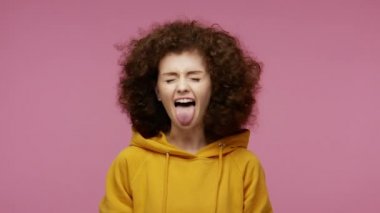 Kaygısız, afro saçlı, kapüşonlu, dilini dışarı çıkaran, kameranın önünde eğlenen, itaatsizliğini ifade eden afro saçlı kız. Stüdyo çekimi pembe arkaplanda izole edildi