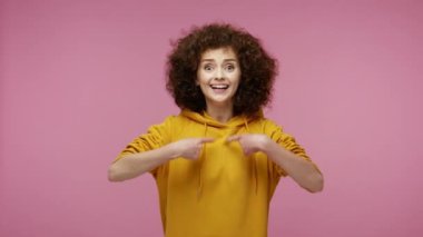Ben buyum! Kapüşonlu mutlu afro saçlı kız kendini gösteriyor, etrafa bakıyor ve başarılarından övünüyor, başarılarından gurur duyuyor. Pembe arka planda kapalı stüdyo çekimleri