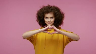 Sevgilim ol! Kapüşonlu güzel, mutlu bir kız saçı. Elleriyle kalp şekli yapıyor ve havayı öpüyor. Aşk sevgisini, romantik hislerini gösteriyor. Pembe arka planda kapalı stüdyo çekimleri