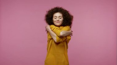 Kapüşonlu sevimli afro saç stili, kendini kucaklayan, kendini beğenmiş ve zevkten gülümseyen narsist egoist duygular, rahatlık konsepti. Stüdyo çekimi pembe arkaplanda izole edildi