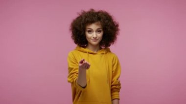 Buraya gel, beni takip et! Kapüşonlu güzel afro saçlı kız tek parmakla işaret ediyor, yaklaşmaya davet ediyor, şakacı cilveli görünüyor. Pembe arka planda kapalı stüdyo çekimleri