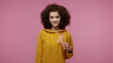 Kapüşonlu, pembe kurdeleli, göğüs kanseri farkındalığının sembolü, kadın sağlığı teşhisi ve hastalık önlemenin sembolü olan güzel afro saçlı bir kız. Pembe arka planda kapalı stüdyo çekimleri