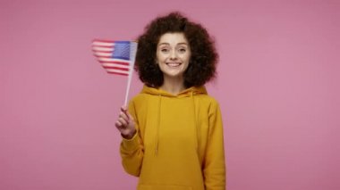 Amerikan bayrağı sallayan, gülümseyen, Bağımsızlık Günü 'nü ulusal bayramını, insan haklarını, demokrasiyi kutlayan kapüşonlu afro saçlı mutlu bir kız. Pembe arka planda kapalı stüdyo çekimleri