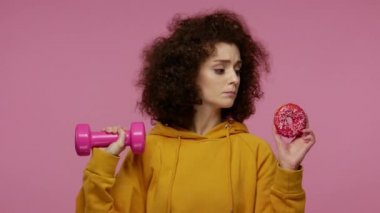 Kapüşonlu kapüşonlu sıska kız saçı, eğitim görüyor ama tatlı çöreğe tutkuyla bakıyor, fitness diyeti vitamini ile abur cubur arasında seçim yapıyor. Stüdyoda çekim izole edildi