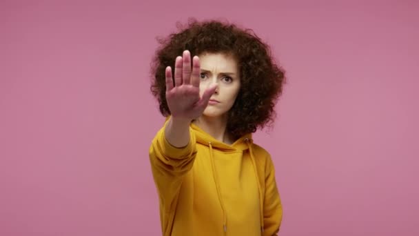 フーディの厳格な積極的な女の子アフロヘアスタイルを停止ジェスチャーを示す 腕を伸ばし 拒絶の概念を 禁止の手の記号を維持する ピンクの背景に隔離された屋内スタジオで — ストック動画