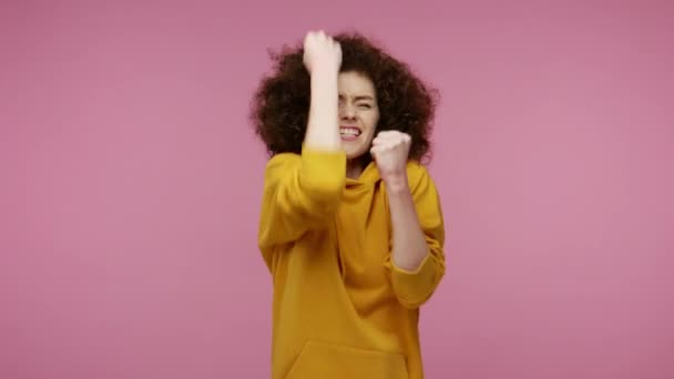 パーカーの叫びで熱狂的な幸せな幸せな女の子アフロヘアスタイル ジェスチャーで拳を上げる私はそれをやった 成功を祝う 勝利と目標達成コンセプト ピンクの背景に隔離されたスタジオショット — ストック動画