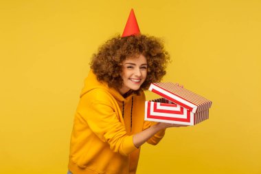 Parti şapkalı komik kıvırcık saçlı kadının portresi kurnaz bir gülümsemeyle kameraya bakıyor ve paketsiz bir hediye kutusu tutuyor, sinsi bir şaka planlıyormuş gibi sırıtıyor. Kapalı stüdyo çekimi, izole.