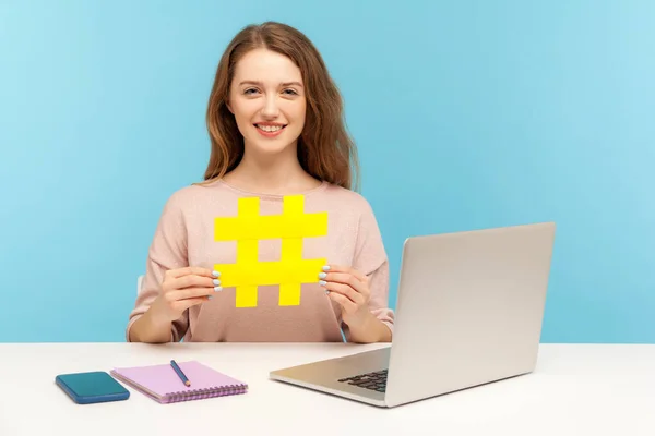 Hash sign. Cheerful attractive woman blogger sitting at workplace with laptop and holding big hashtag symbol, sharing viral content, tagged message. indoor studio shot isolated on blue background