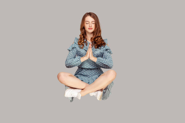 Парит в воздухе. Спокойная, спокойная, расслабленная девушка взъерошить платье левитации с молитвенным жестом, держать глаза закрытыми, медитировать сидя в положении йоги. Крытая студия, снятая на сером фоне
