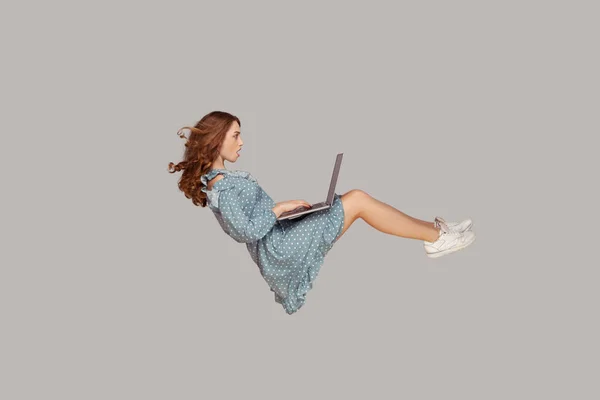 空気中でホバリング 驚きの少女ラッフルドレスを持ち上げ ラップトップの画面を見て 空中を飛んでいる間 Webソーシャルネットワークをサーフィン驚きました グレーの背景に隔離された屋内スタジオで — ストック写真