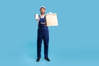 Mavi üniformalı gülümseyen profesyonel kurye alışveriş poşeti taşıyor ve reklam görüntüsü için cep telefonunu gösteriyor. Yemek dağıtım servisi uygulaması. Stüdyo çekimi izole