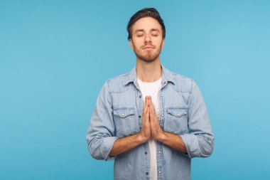 Rahatlama, huzur dolu bir zihin. Çalışan kot gömlekli sakin bir adamın portresi kapalı gözler ve dua jesti, meditasyon, konsantre düşünceler. Mavi arka planda kapalı stüdyo çekimleri