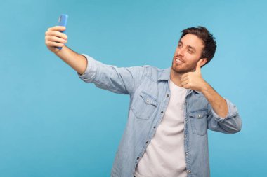 Kot gömlekli olumlu yakışıklı bir adamın portresi video görüşmeleri ve işaret dili gibi işaretler, cep telefonuyla online konuşma, selfie çekme gibi. Stüdyo çekimi, mavi arkaplan