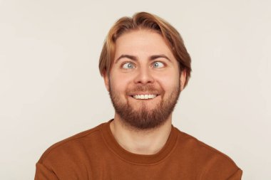 Closeup portrait of idiot, dumb bearded man looking cross-eyed with stupid smile, fooling around, making silly face, brainless comical expression. indoor studio shot isolated on gray background